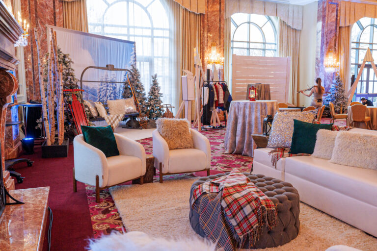 A wide-angle view of a winter lodge-themed event space with cozy seating, faux snow-covered trees, a ski lift photo op, and a violinist performing in the background.