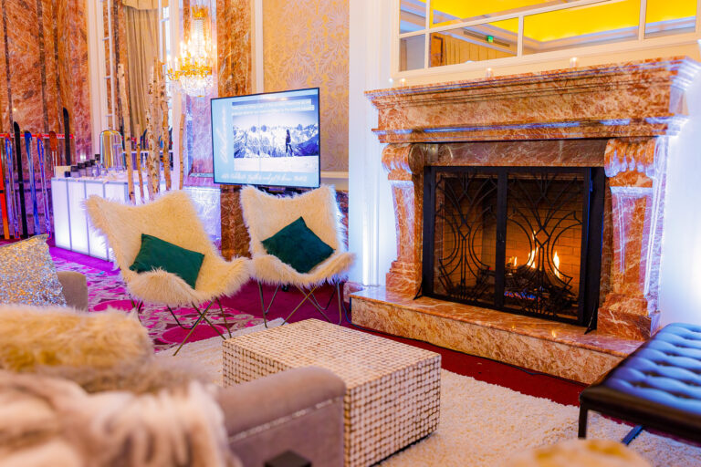 A cozy fireplace lounge setup with faux fur chairs, textured pillows, a digital screen, and glowing ski-themed decor in a grand indoor venue.