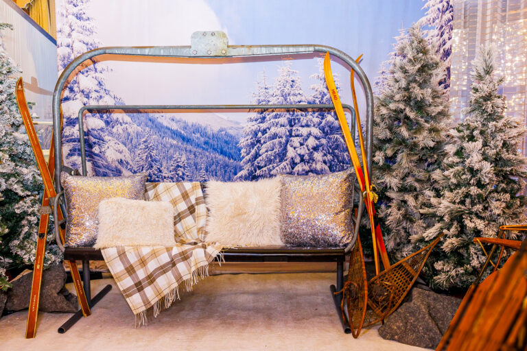 A cozy winter-themed lounge area featuring a vintage ski lift bench with faux fur and plaid pillows, surrounded by frosted trees, antique skis, and snowshoes.