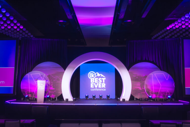 A wide view of the Best Ever Conference stage with circular backdrop and striking LED stage lighting.