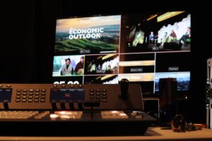 A close-up of a video switcher in front of a screen showing a live broadcast feed for the Economic Outlook Summit, with multiple camera angles and branding visible. Professional AV equipment rental setup at business event