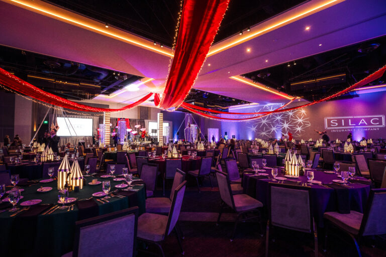 Gala ballroom with red ceiling drapery, striped tent centerpieces, and stage lighting at SILAC Insurance event