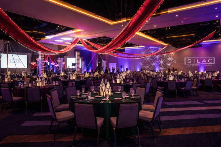 Wide view of banquet hall with red ceiling drapery, themed centerpieces, and branded lighting at the SILAC Insurance Gala 2024