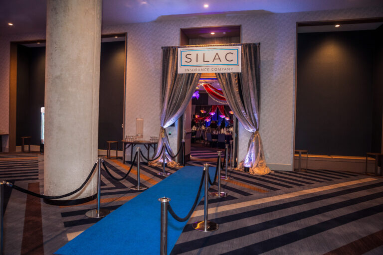 Event entry with silver drapery, branded signage, and blue carpet walkway at the SILAC Insurance Gala 2024