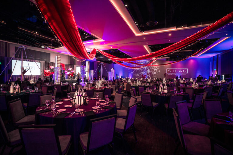 Wide view of gala setup with red ceiling drapery, striped centerpieces, stage lighting, and projection at SILAC Insurance Gala 2024