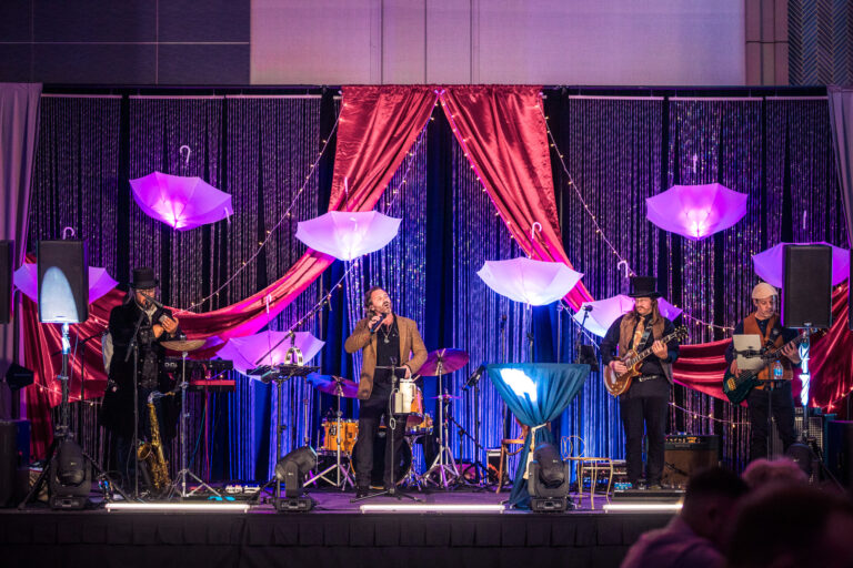 Live band performs on a decorated stage with umbrella props, drapery, and ambient lighting at a gala event
