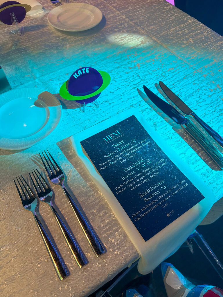 Close-up of a glowing blue place setting with a cosmic-themed menu and custom Saturn name card reading “Kate” at Access DMC’s space-themed dinner by In The Event.