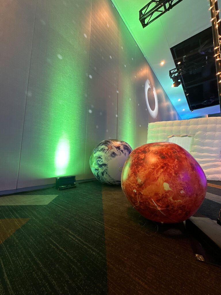 Oversized inflatable planets illuminated by colorful green and blue uplighting in a space-themed event setup at the Grand Hyatt Salt Lake City by In The Event.