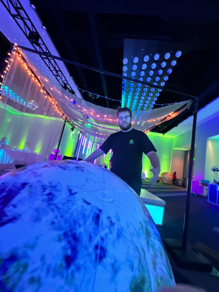 Event technician setting up a glowing planetary prop under LED canopy lighting with colorful uplighting and string lights during Access DMC’s space-themed event at the Grand Hyatt Salt Lake City.