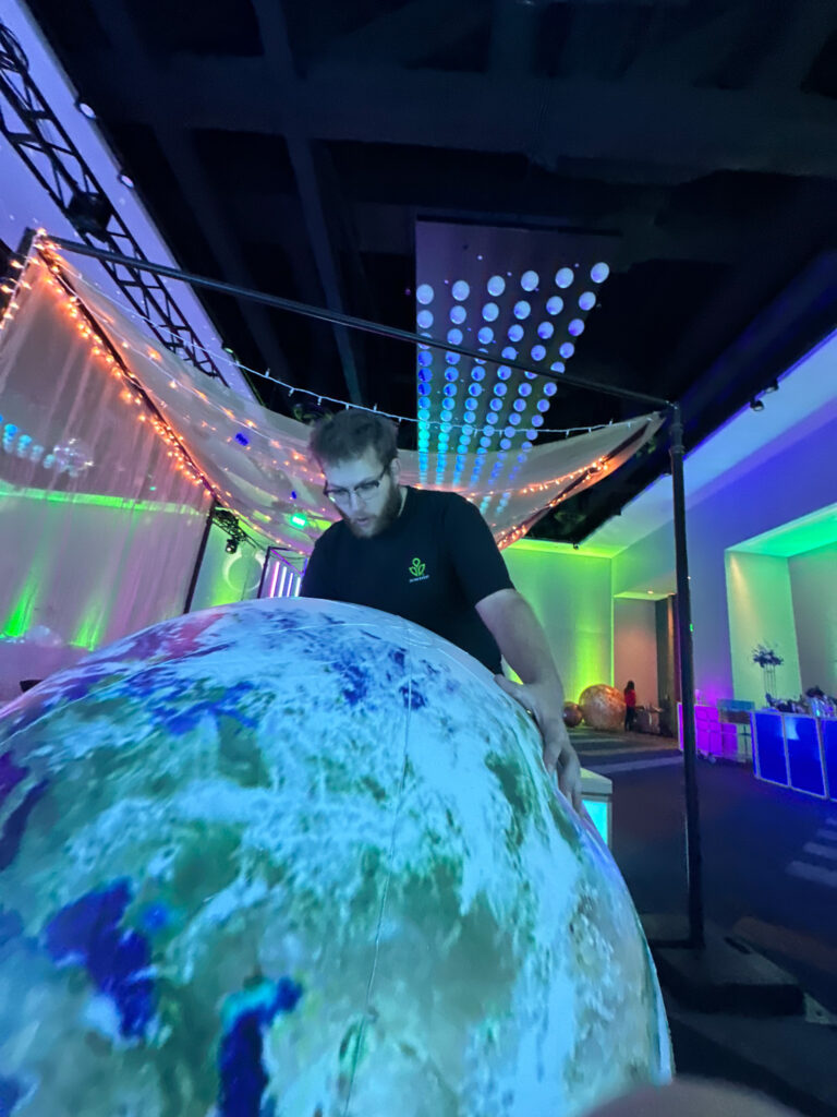 Event technician adjusts a large inflatable Earth beneath glowing LED canopy lights during the Access DMC space-themed event at the Grand Hyatt Salt Lake City.