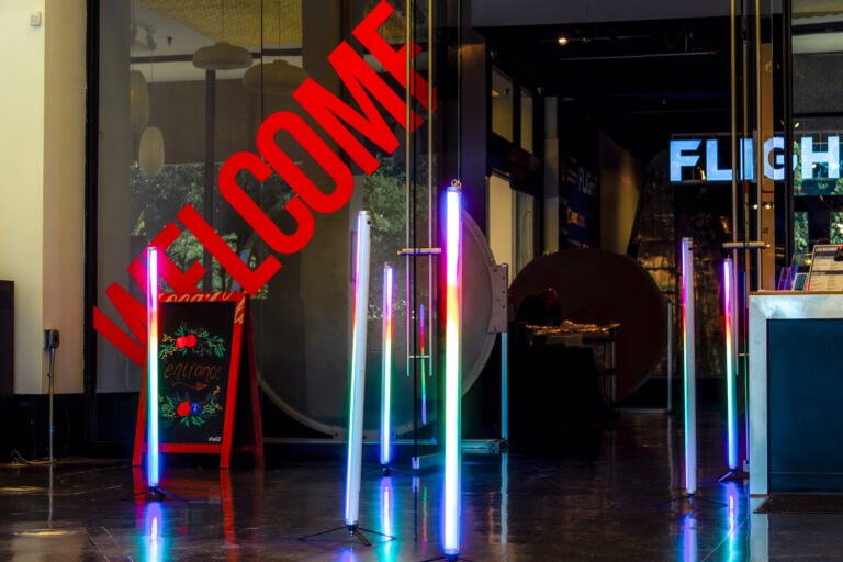 Colorful LED tube lights line an event entryway under a bold red “WELCOME” sign, creating a modern illuminated pathway.
