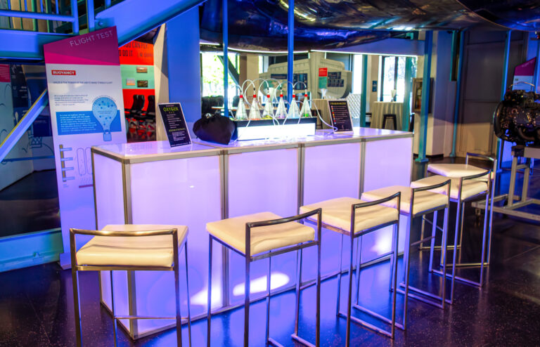 Modern LED-lit bar setup with white modular panels, sleek metal barstools, and illuminated countertop in a science-themed venue.