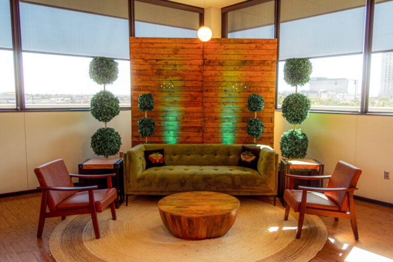 Mid-century lounge setup featuring a green velvet sofa, leather armchairs, wooden coffee table, and topiary accents against a wood panel backdrop.