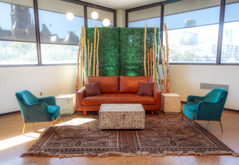 Modern lounge setup with a brown leather sofa, teal velvet chairs, birch wood accents, and a boxwood hedge backdrop.