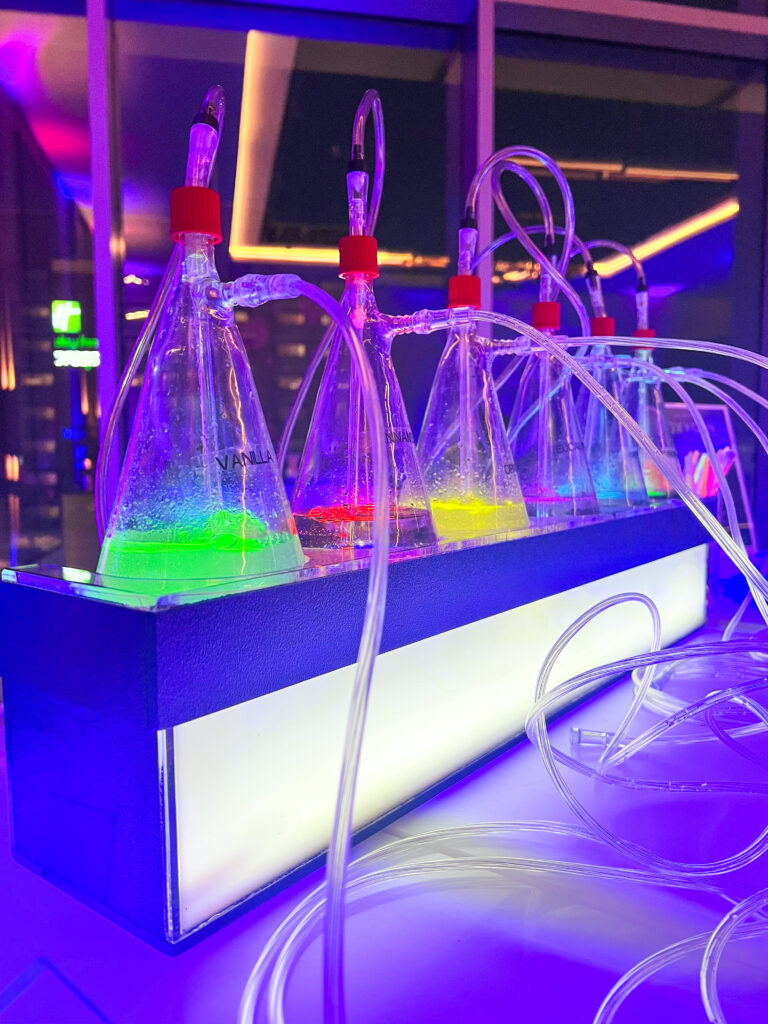 Close-up of a laboratory-themed drink dispenser featuring scientific flasks with brightly colored liquids, tubes, and a base illuminated by LED furniture rentals lights.