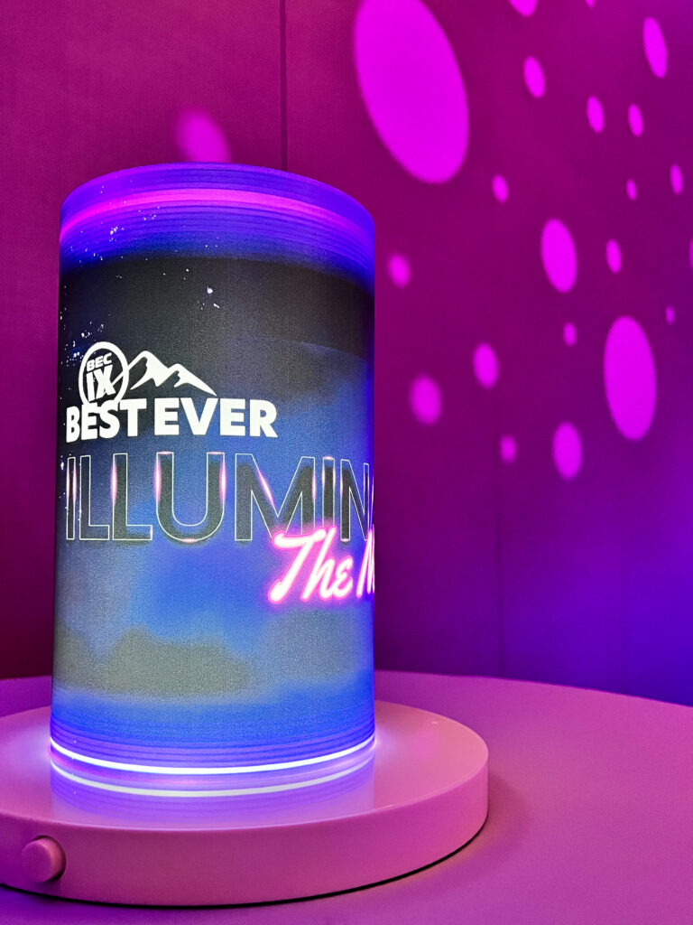 Close-up of a glowing cylindrical LED centerpiece rentals with custom event signage branding, set on a white table under pink and purple custom lighting solutions.