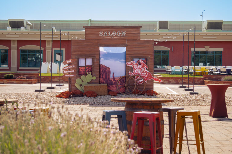 Outdoor event featuring a custom Western Saloon facade and themed event props like wagon wheels, a cactus, and wood barrel tables.