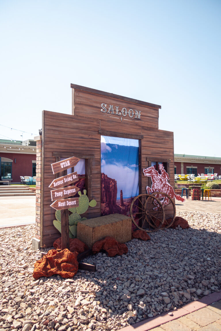 Close-up of a custom Western Saloon facade event backdrop with directional custom event signage, large desert event props like wagon wheels, and faux cacti.