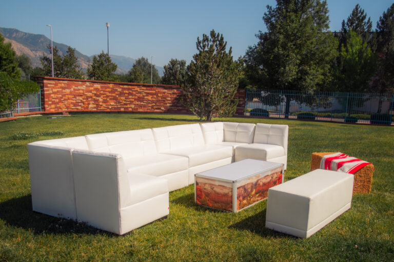 White sectional event lounge furniture and ottoman on a bright green lawn with a custom-branded coffee table, mountains in the background, and a rustic hay bale.