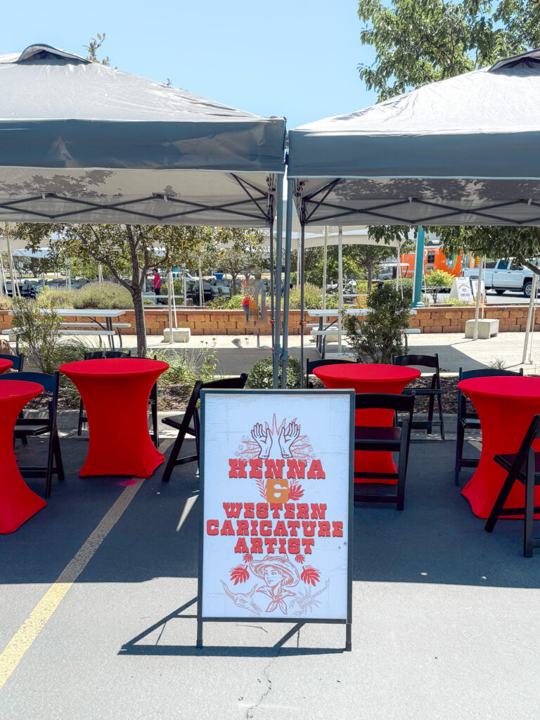 Outdoor event area with gray pop-up tents, red cocktail tables, black chairs, and A-frame custom event signage advertising a henna and western caricature artist.