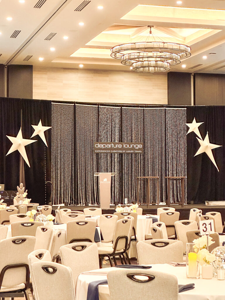 Elegant ballroom setup with draped event backdrops, a textured stage design, large white star event props, lighting rentals, and formal table decor.