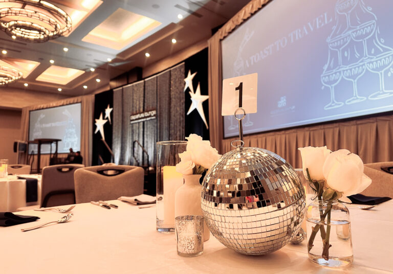 Elegant event table setting with a disco ball centerpiece rentals, white roses, candles, and a table number, with a large screen displaying event branding in the background.