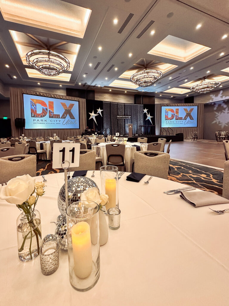 Wide view of a gala dinner setup with round tables, disco ball centerpiece rentals, custom-branded screens for video rentals, a central stage rentals area, and tiered chandelier lighting rentals.