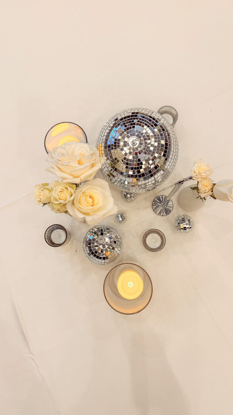 Overhead view of elegant table decor featuring mirrored disco balls in various sizes, soft yellow roses, and lit candles in glass holders for centerpiece rentals.