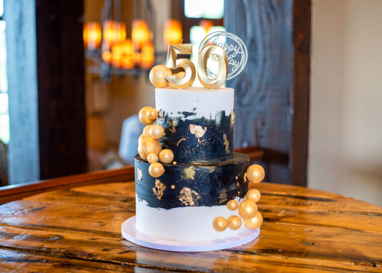 Elegant two-tier black and white cake with gold accents and “50” topper displayed on a rustic wooden table at a birthday celebration.