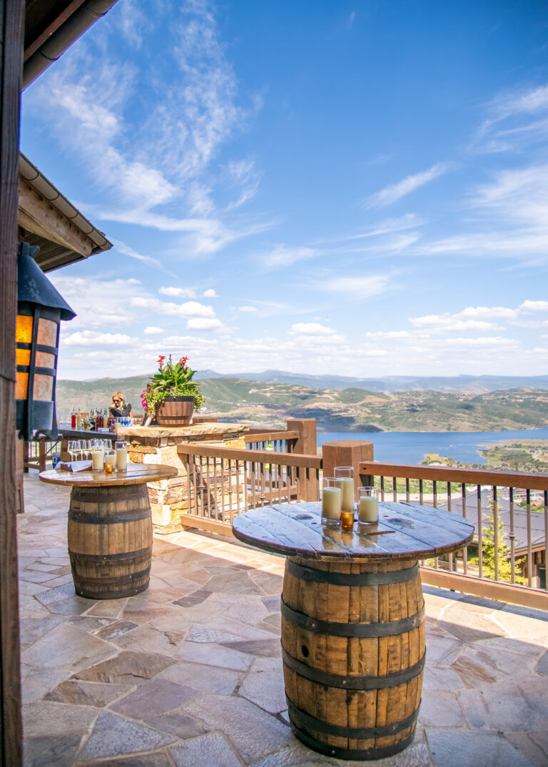 Rustic patio setup overlooking a mountain lake, featuring wooden barrel cocktail tables decorated with candles and surrounded by scenic views.
