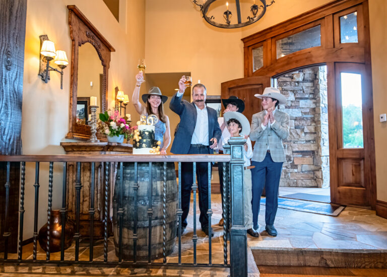Family celebrating indoors with cowboy hats, floral arrangements, and a black and gold cake displayed on a rustic barrel table at a Western-themed event.