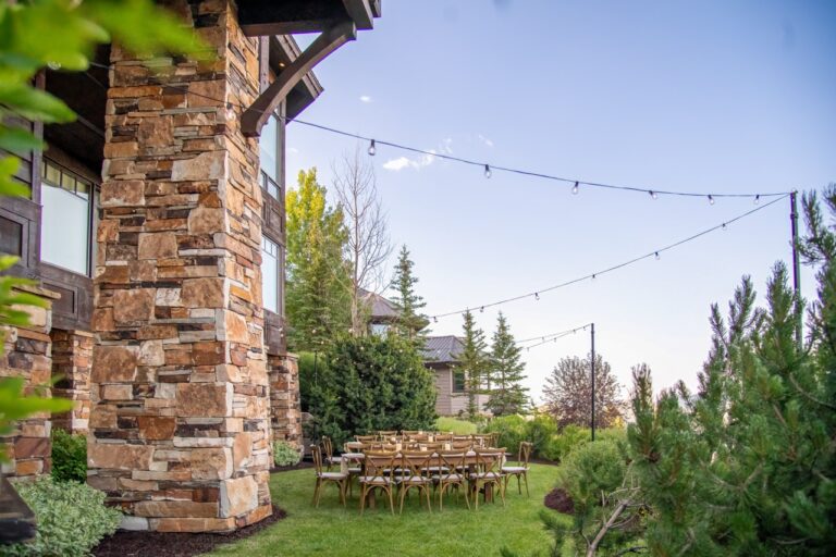 Charming outdoor event setup featuring wooden crossback chairs and farm tables beneath hanging string lights beside a stone mountain lodge.
