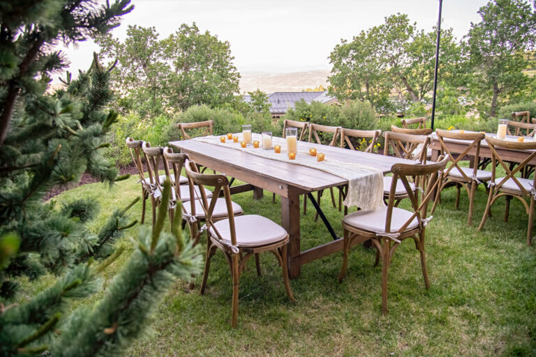 Outdoor event setup featuring a long wooden farm table with crossback chairs, candle décor, and neutral table runner surrounded by lush greenery.
