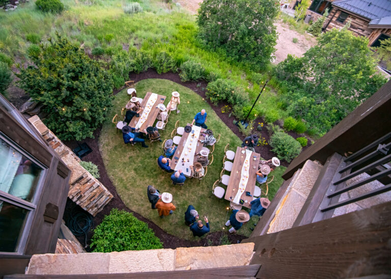 Overhead view of guests seated at long wooden farm tables on a grassy lawn, surrounded by lush greenery and string lights for a rustic Western-themed dinner.