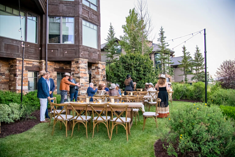 Guests gather for a rustic outdoor dinner with wooden farm tables, crossback chairs, and string lights at a mountain home event, styled in a Western theme.