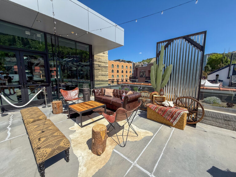 Outdoor Western-themed lounge featuring leather seating, rustic wood tables, a corrugated metal backdrop, cactus prop, and hay bale décor under string lights.