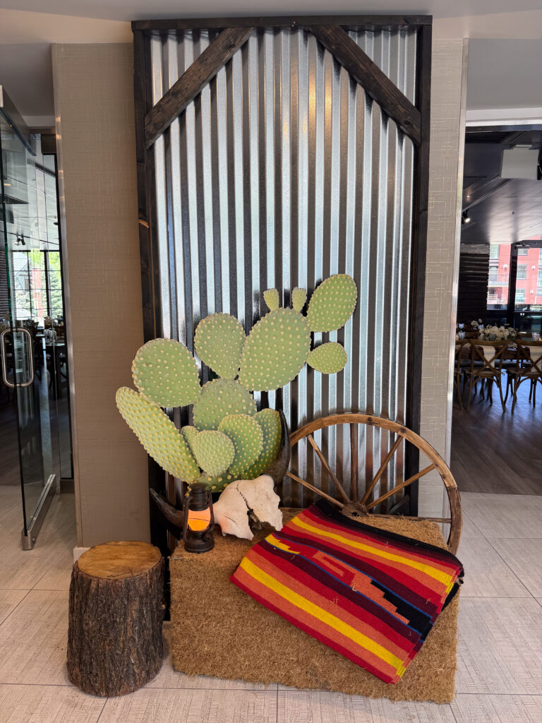 Rustic Western display featuring a prickly pear cactus prop, wagon wheel, hay bale, and colorful striped blanket with skull and lantern accents.
