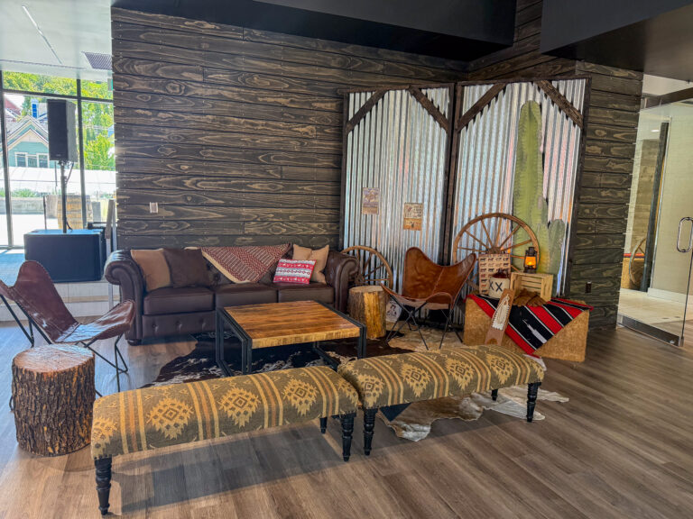 Rustic Western lounge featuring a leather butterfly chair, hay bale covered with a red patterned blanket, wagon wheels, and a cactus backdrop with lantern lighting.
