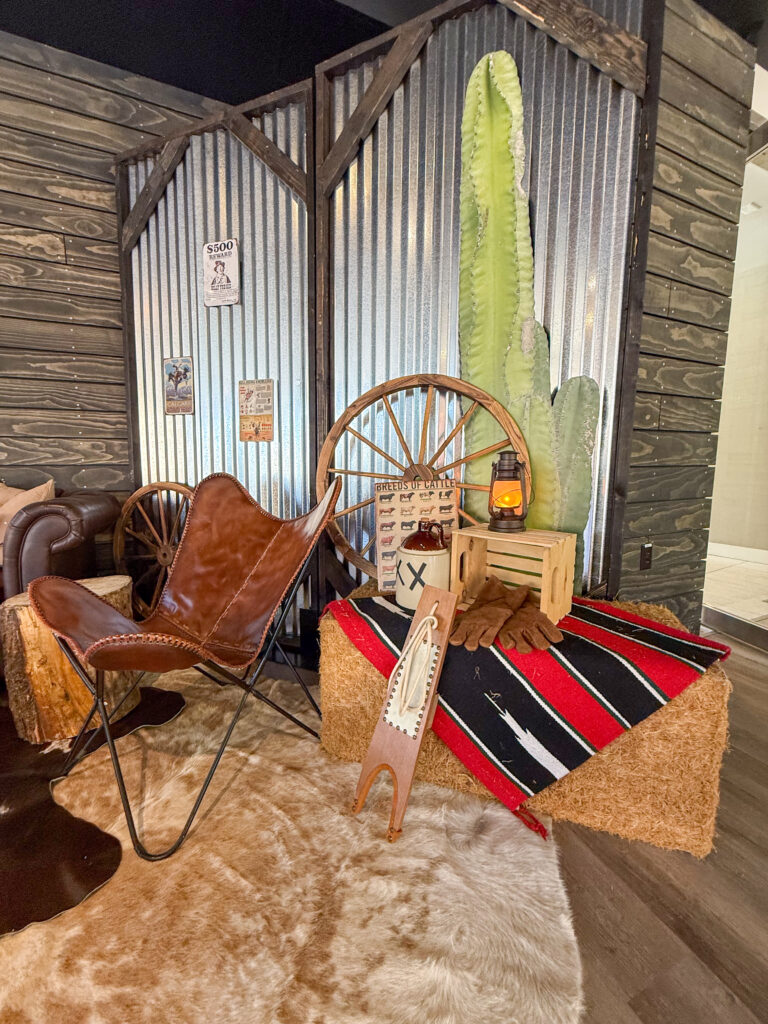 Rustic Western lounge featuring a leather butterfly chair, hay bale covered with a red patterned blanket, wagon wheels, and a cactus backdrop with lantern lighting.