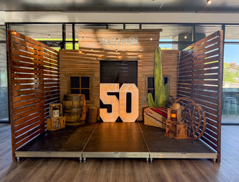 Indoor Western-themed saloon setup featuring a light-up “50” marquee, cactus prop, wagon wheel décor, and rustic barrel cocktail table with floral accents.