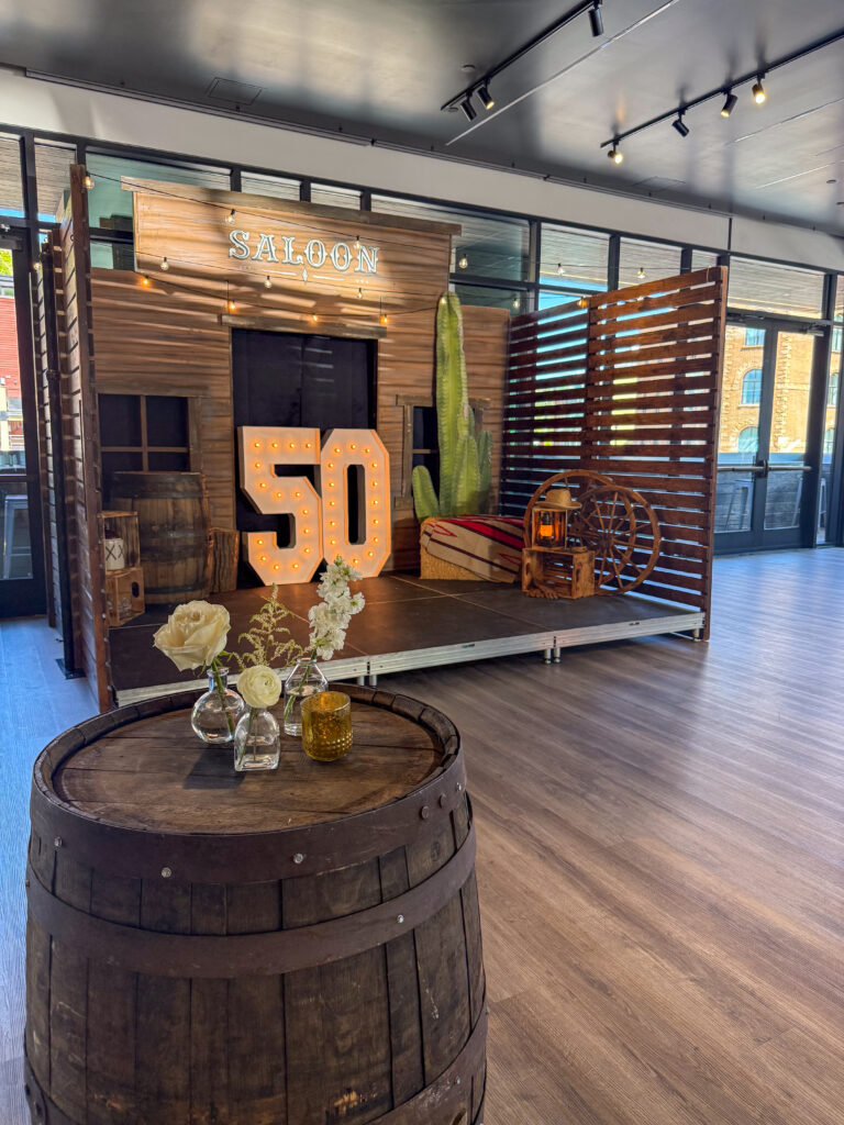 Indoor Western-themed saloon setup featuring a light-up “50” marquee, cactus prop, wagon wheel décor, and rustic barrel cocktail table with floral accents.