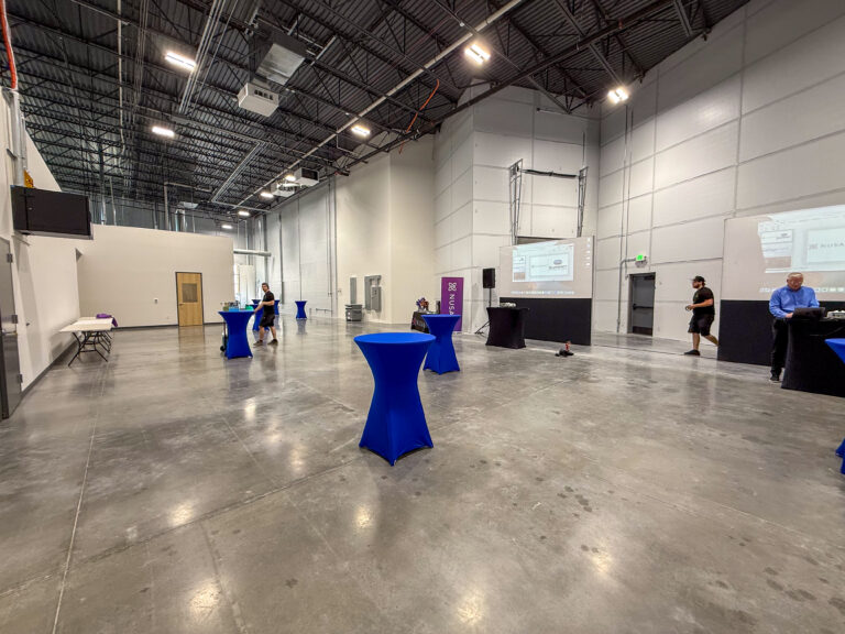 Large warehouse-style venue prepared for a corporate event with blue spandex cocktail tables, projection screens, and branded signage.