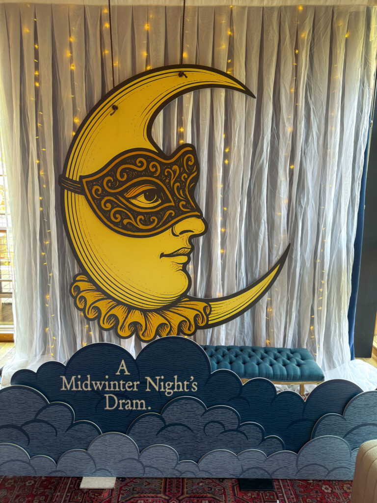 Golden crescent moon with ornate mask and “A Midwinter Night’s Dram” lettering against a draped backdrop with string lights