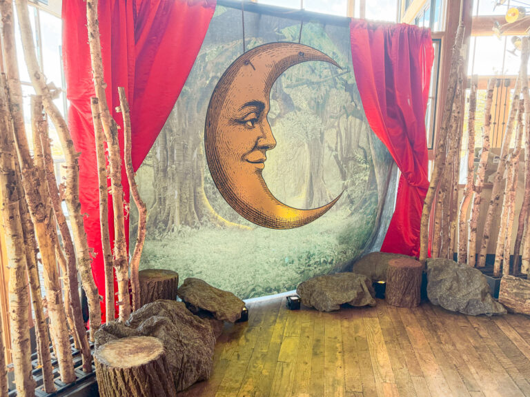 Celestial-themed photo backdrop featuring a vintage crescent moon illustration framed by red velvet drapes and forest-inspired props at the High West Distillery Party