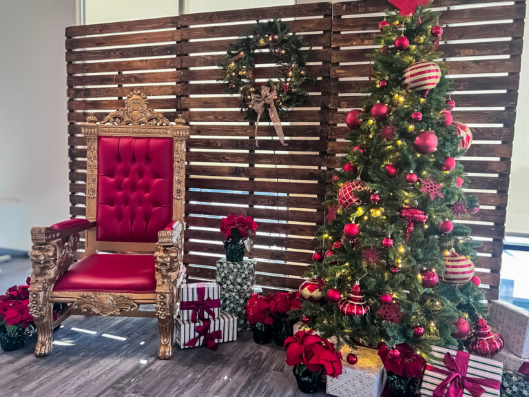 Santa throne with red upholstery and gold detailing beside a decorated Christmas tree, gifts, wreath, and poinsettias at Trucordia event.