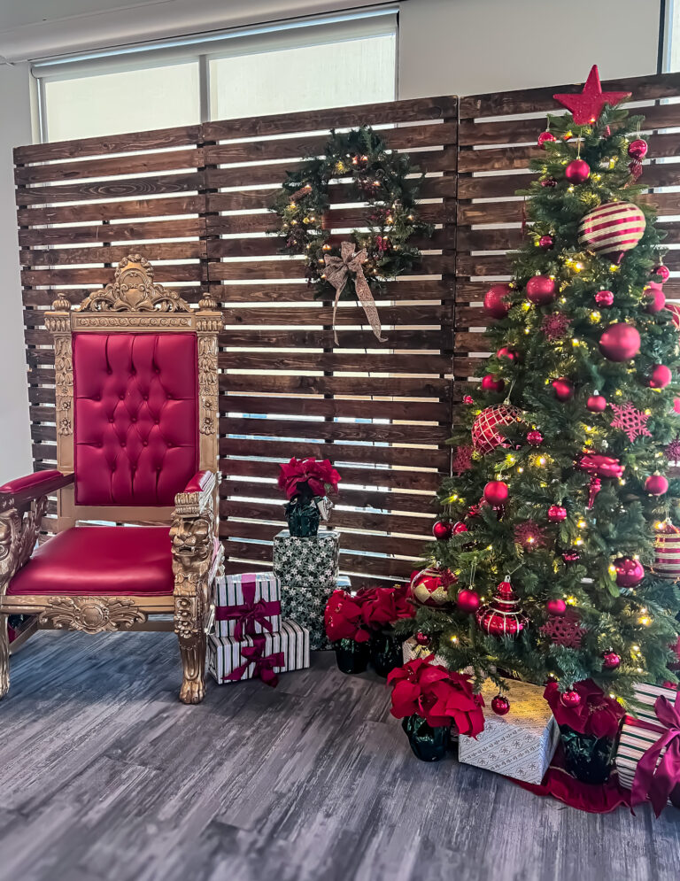 Santa throne with red tufted upholstery beside decorated Christmas tree, wrapped presents, poinsettias, and wreath at Trucordia holiday setup.