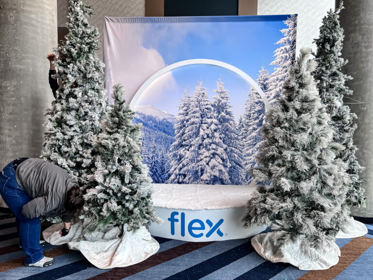 Team member setting up a snow globe photo backdrop with flocked Christmas trees and the Flex logo at the Flex Holiday Party.