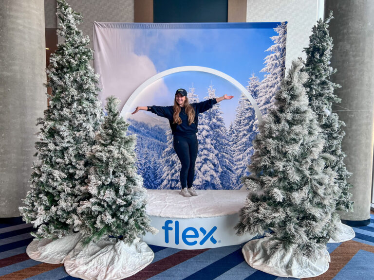 Woman posing inside a snow globe-style photo booth surrounded by flocked Christmas trees at the Flex Holiday Party.