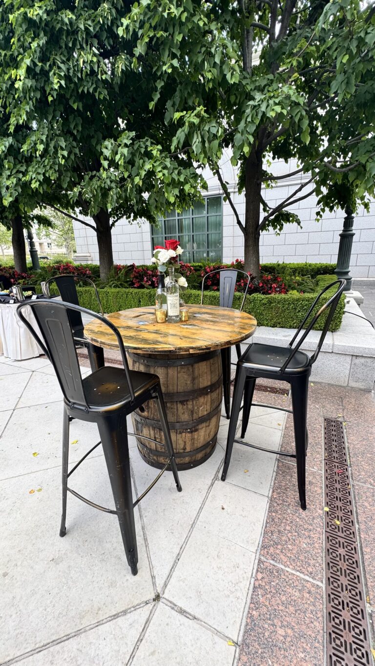 Outdoor event setup featuring a rustic wooden barrel table with black metal chairs and floral centerpiece, perfect for cocktail hour or garden gatherings.