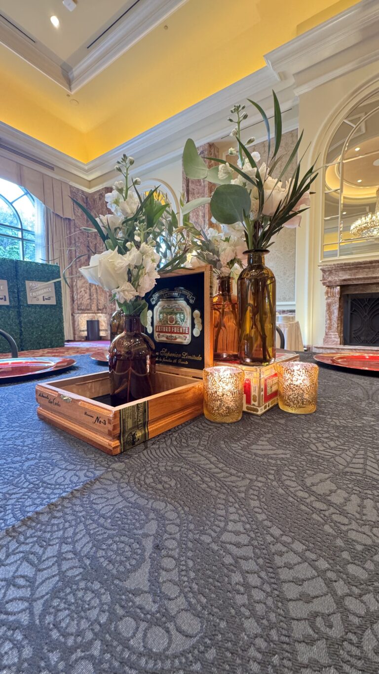 Table centerpiece featuring amber glass vases, white florals, gold votive candles, and an open cigar box accent on a textured black linen table.